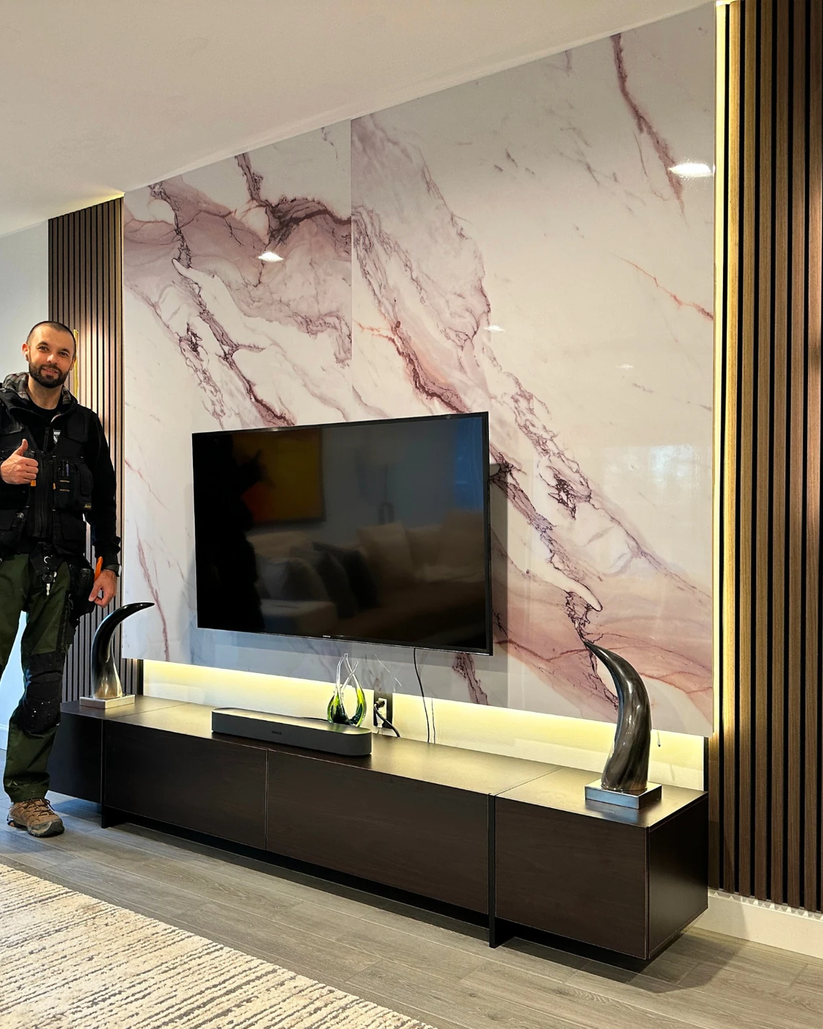 Roman beside a completed marble and wood slat statement media wall with sconces and mounted TV