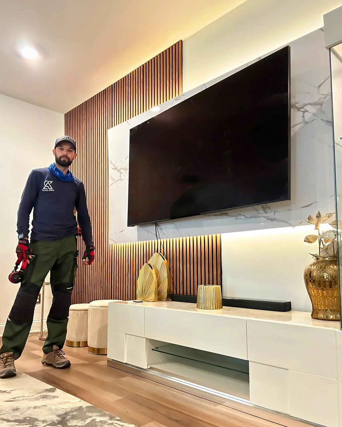 Roman beside a completed mixed-material media wall with wood slats, marble-look paneling, LED lighting, and TV