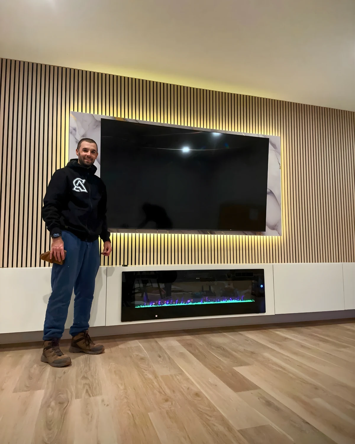 Roman beside a completed full-width acoustic slat media wall with backlit TV panel, electric fireplace, and custom cabinetry
